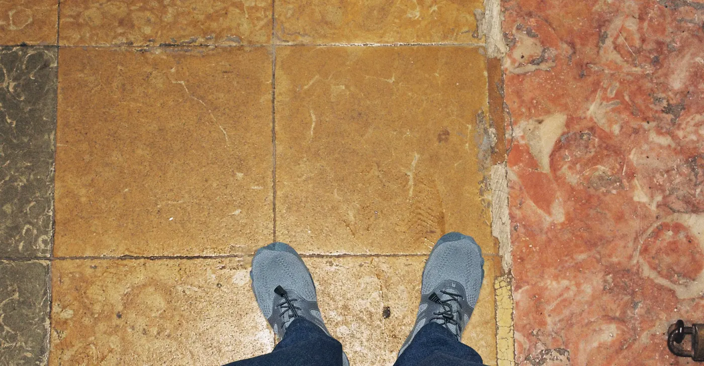 Looking down at worn yellow limestone floor tiles beside a pink marble panel, with the designer’s shoes at the bottom edge