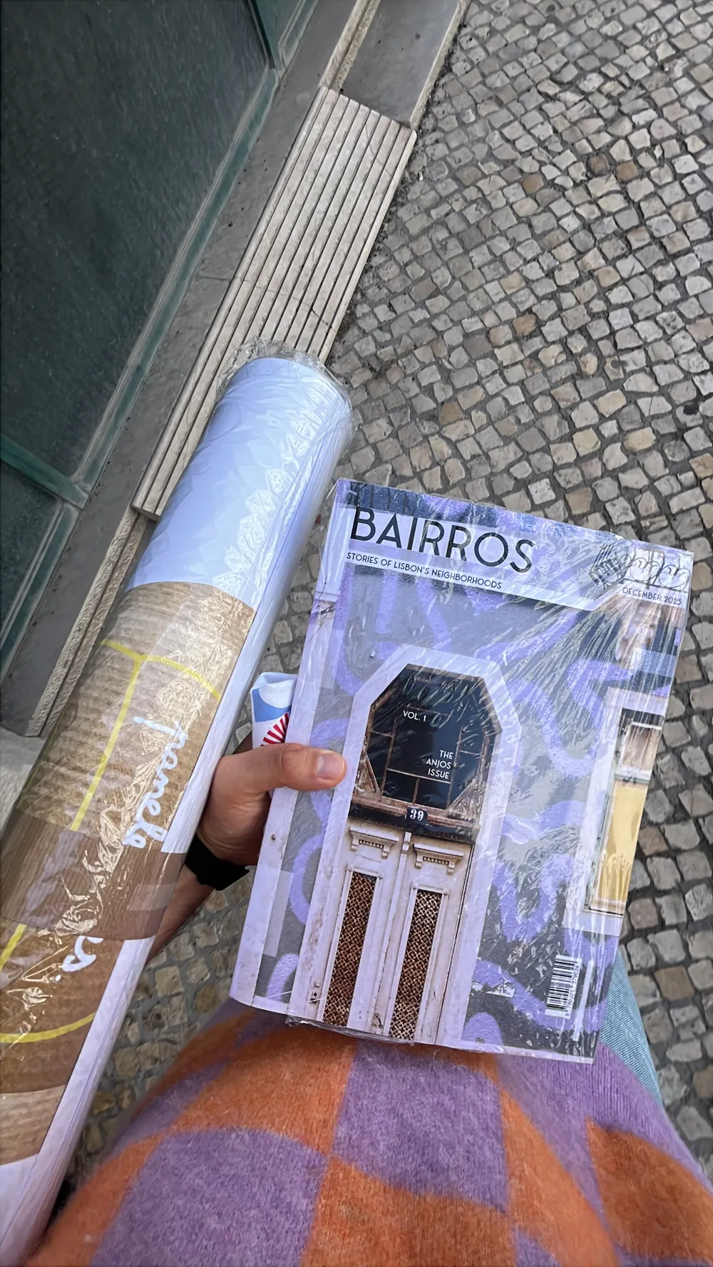 A hand holding the printed, shrink-wrapped Bairros magazine beside a rolled-up Paneleirices poster, over a purple and orange checked sweater, on a cobbled pavement