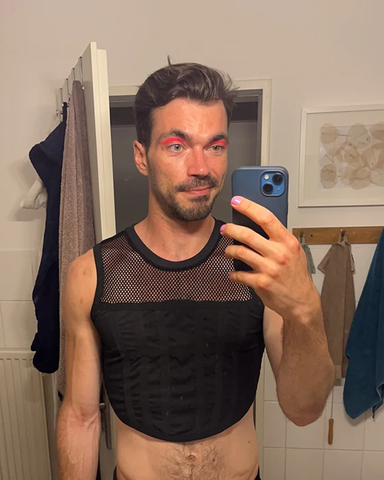 A man in a black mesh top with pink eye makeup, taking a mirror selfie