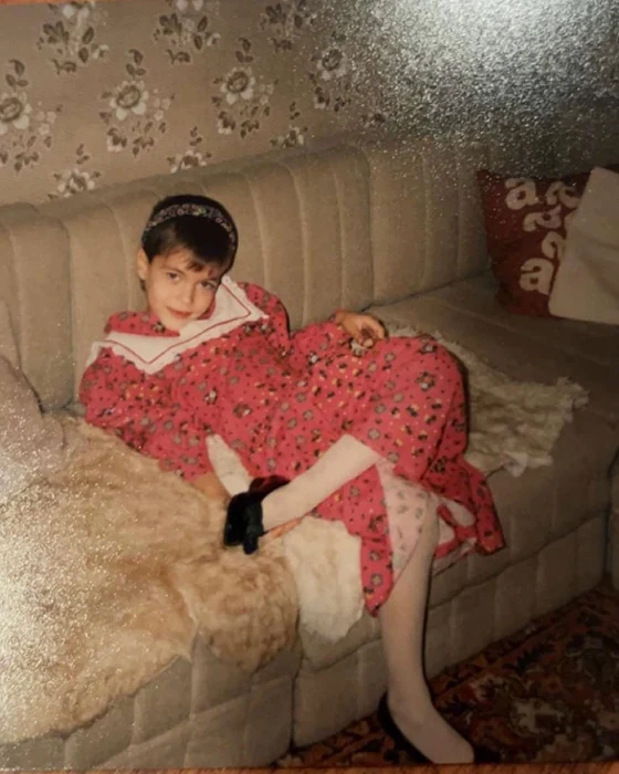 A childhood photo of a boy in a red floral dress, sitting on a sofa