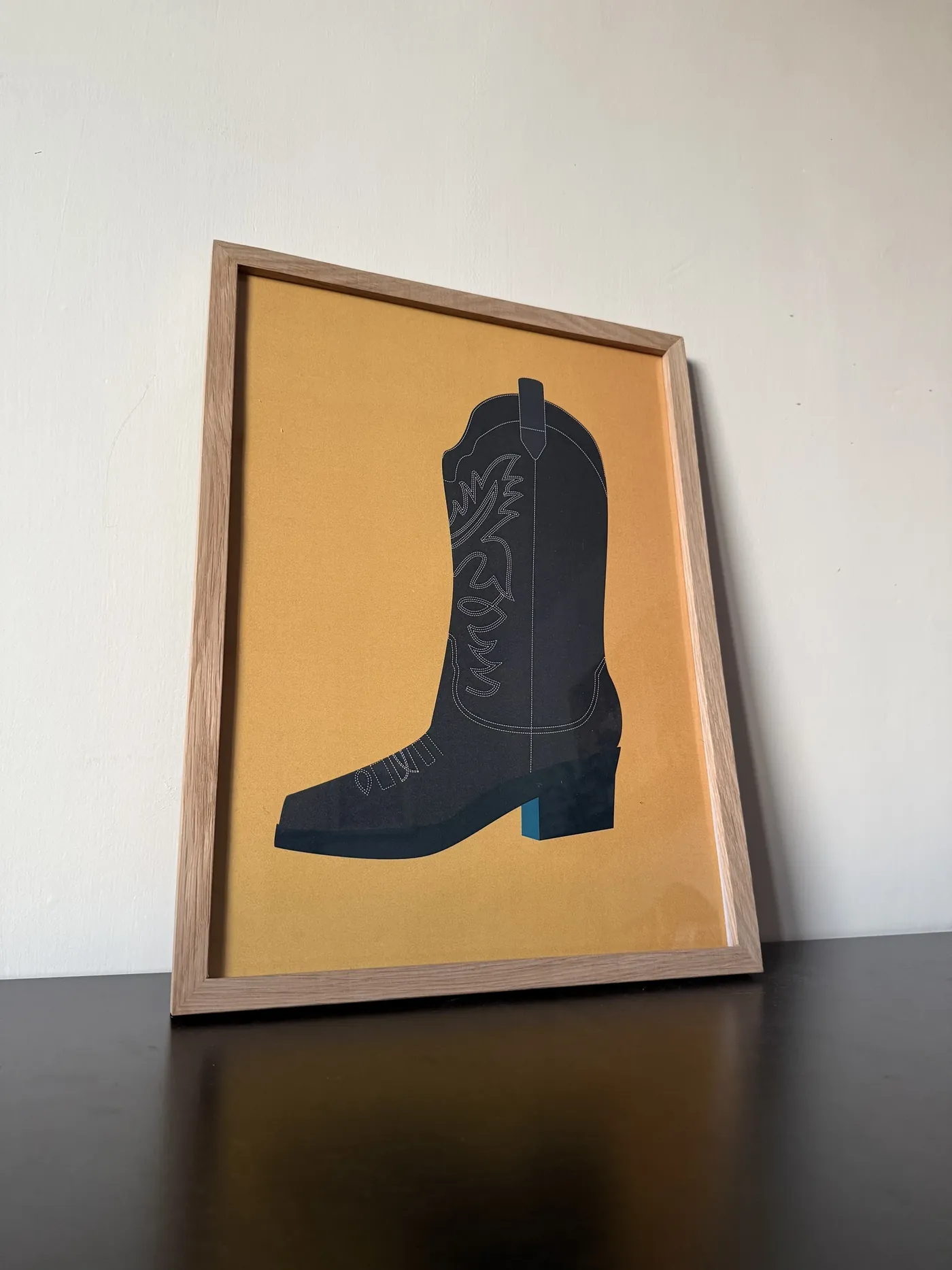 The printed boot poster in an oak frame, leaning against a white wall on a dark table
