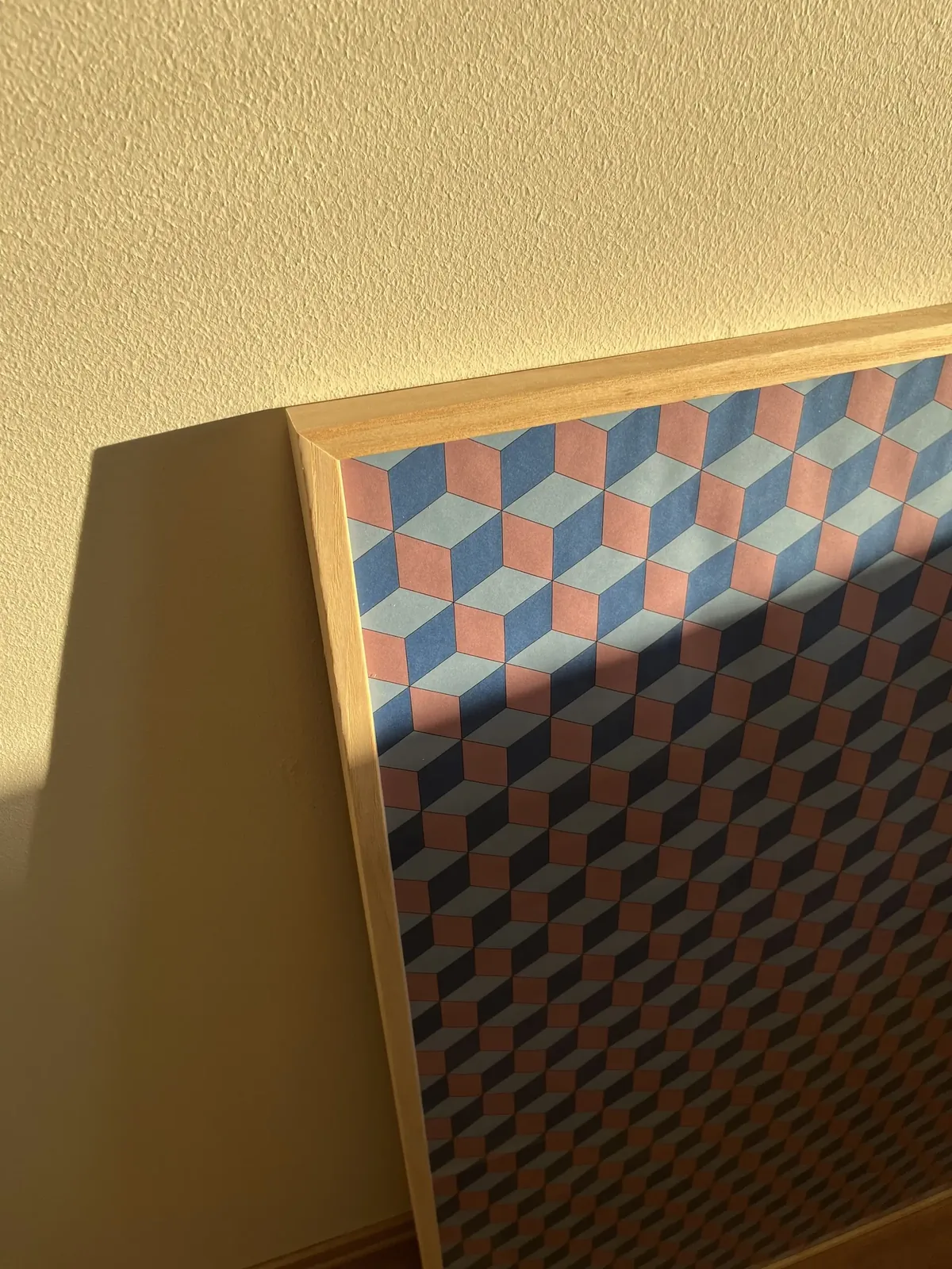 Close-up of the framed rhombille poster in warm sunlight, the frame casting a shadow across the pattern
