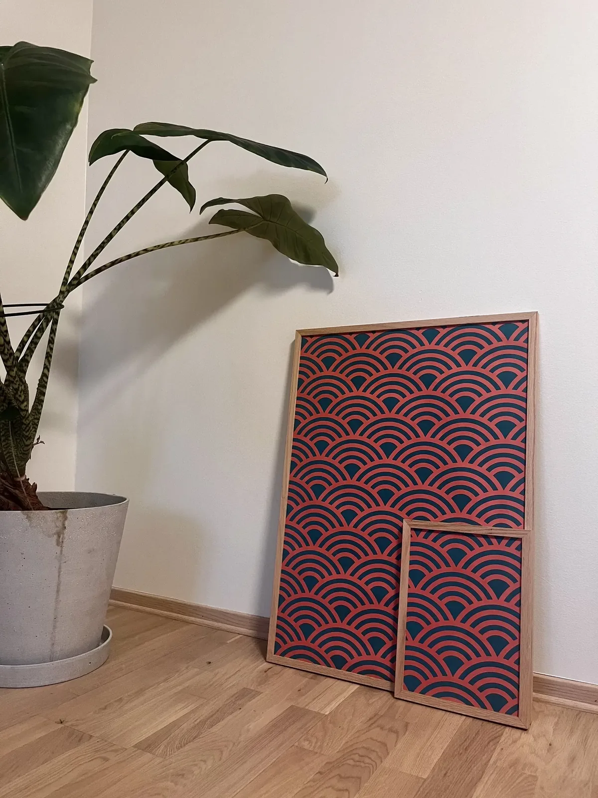 Two framed seigaiha posters in orange-red and dark teal, a small one in front of a large one, leaning against a wall next to a plant