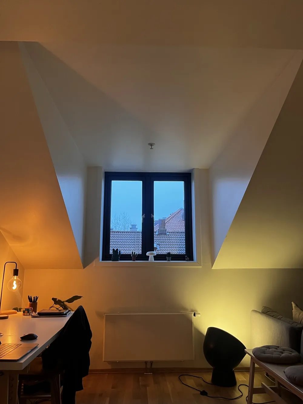 A warm attic room at dusk with a dormer window and a lamp glowing yellow, the view the illustration is based on