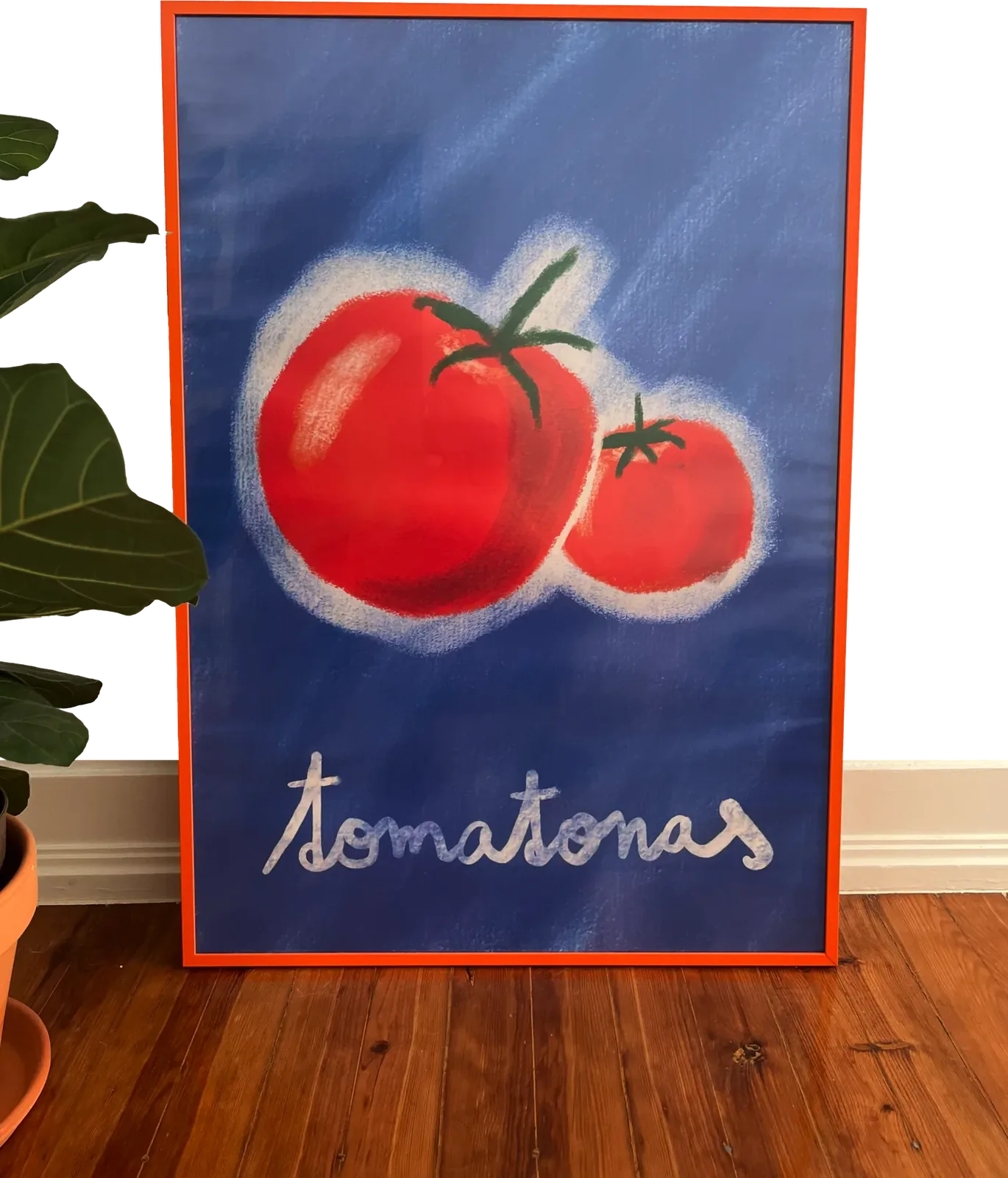 The tomatonas poster in an orange frame, leaning against a wall next to a plant: two red tomatoes on a blue background with the word tomatonas in chalky white handwriting