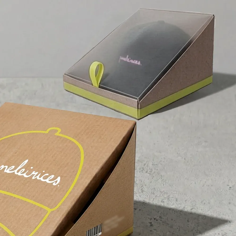 A kraft display box with a clear lid and a neon pull tab holding a black cap, above the corner of a second kraft box printed with a yellow line drawing of a cap